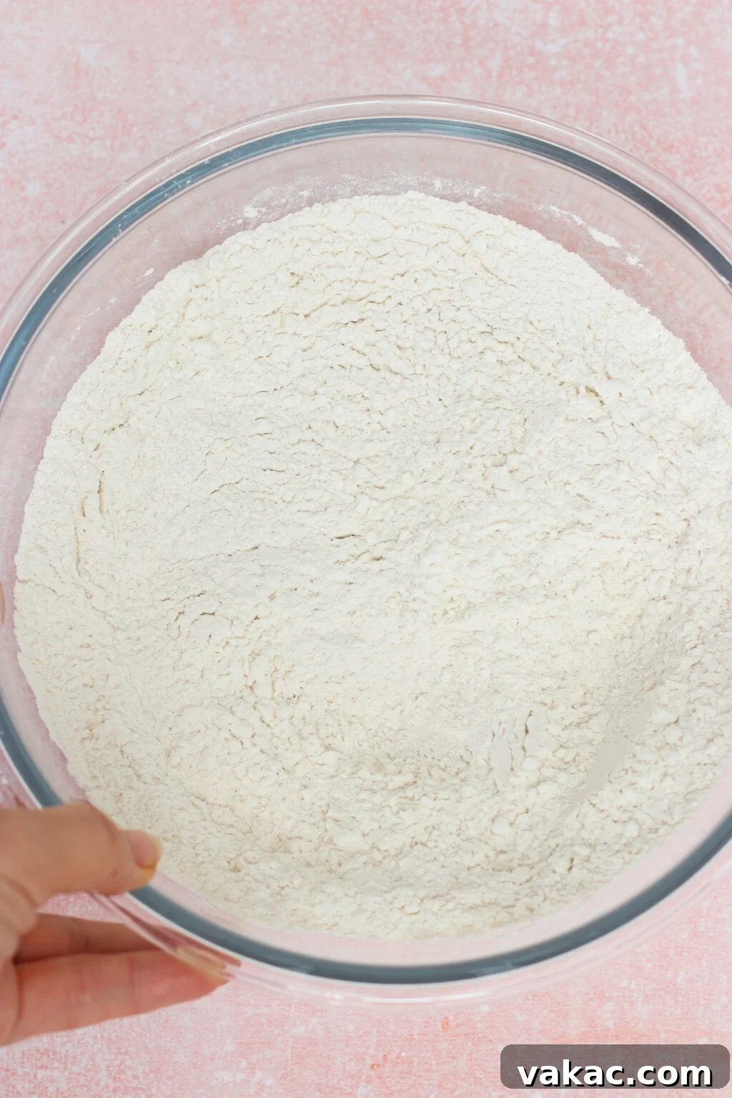 High-Protein Cottage Cheese Bagels Recipe for Homemade Bagels 4 A hand holding a clear glass bowl filled with white flour, set on a light pink textured surface.