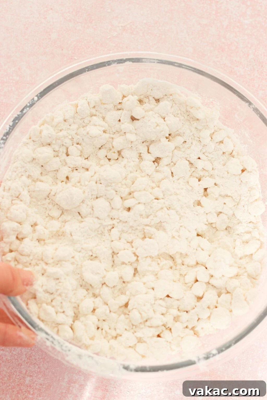 High-Protein Cottage Cheese Bagels Recipe for Homemade Bagels 5 A clear glass bowl filled with a coarse, crumbly mixture of flour and butter sits on a pale pink surface. A hand is holding the edge of the bowl.