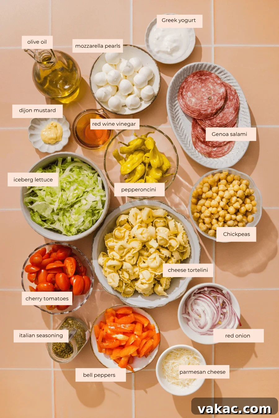 A colorful spread of ingredients in bowls on a tiled surface, including olive oil, mozzarella pearls, Greek yogurt, Genoa salami, lettuce, pepperoncini, chickpeas, tortellini, tomatoes, bell peppers, onion, and cheese.