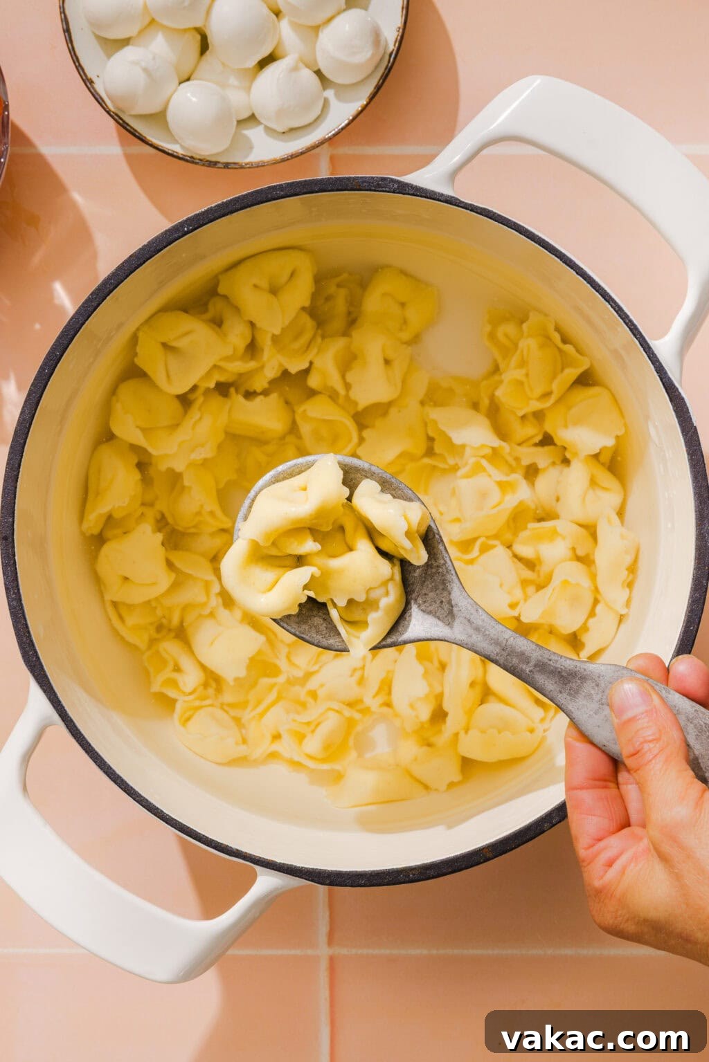 A hand holds a large spoon, scooping cooked cheese tortellini from a white pot. In the background, a plate with small mozzarella balls is visible on a light pink tiled surface.