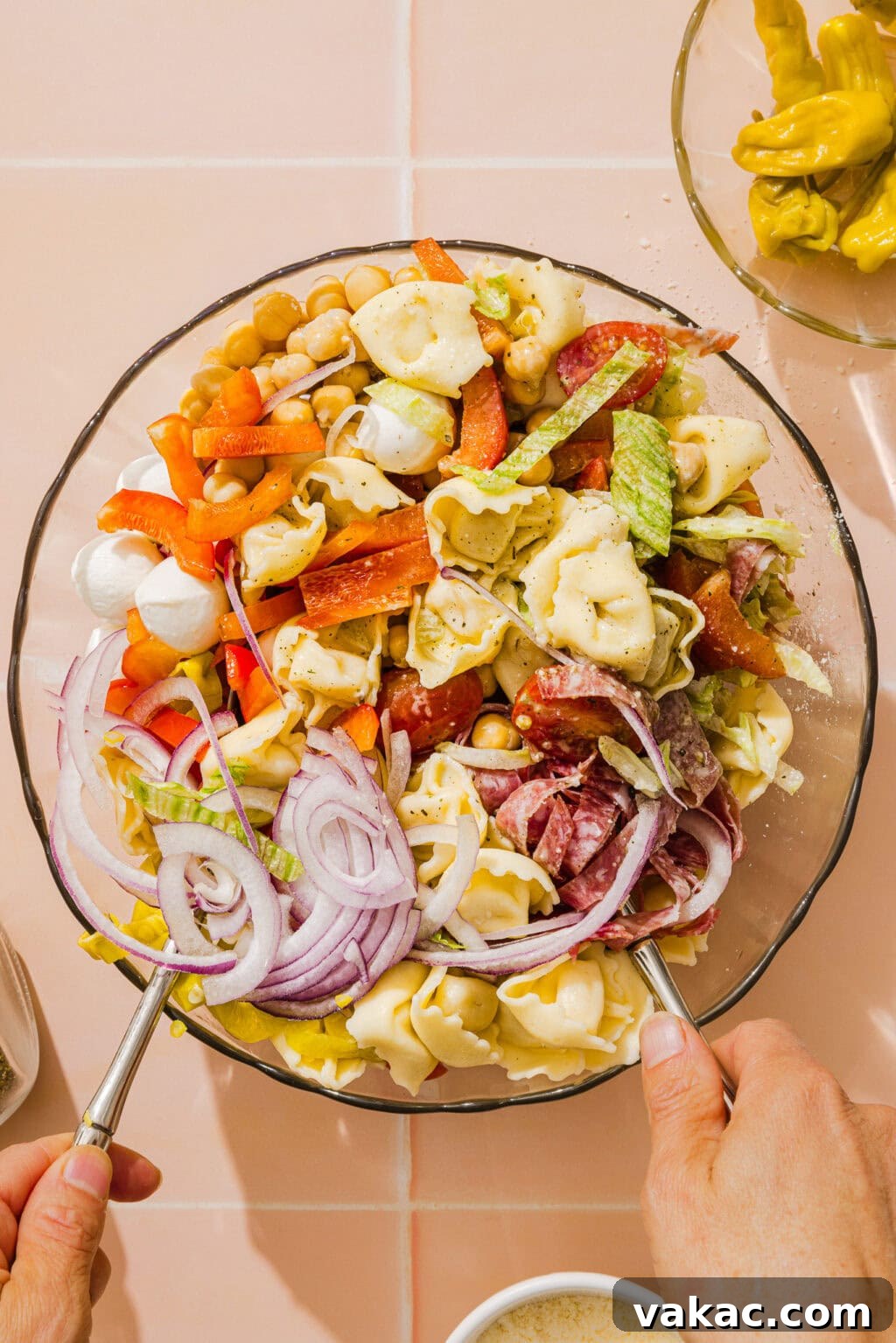A person tosses a large bowl of pasta salad with tortellini, sliced red onions, bell peppers, salami, chickpeas, mozzarella balls, celery, and carrots, using two serving spoons.
