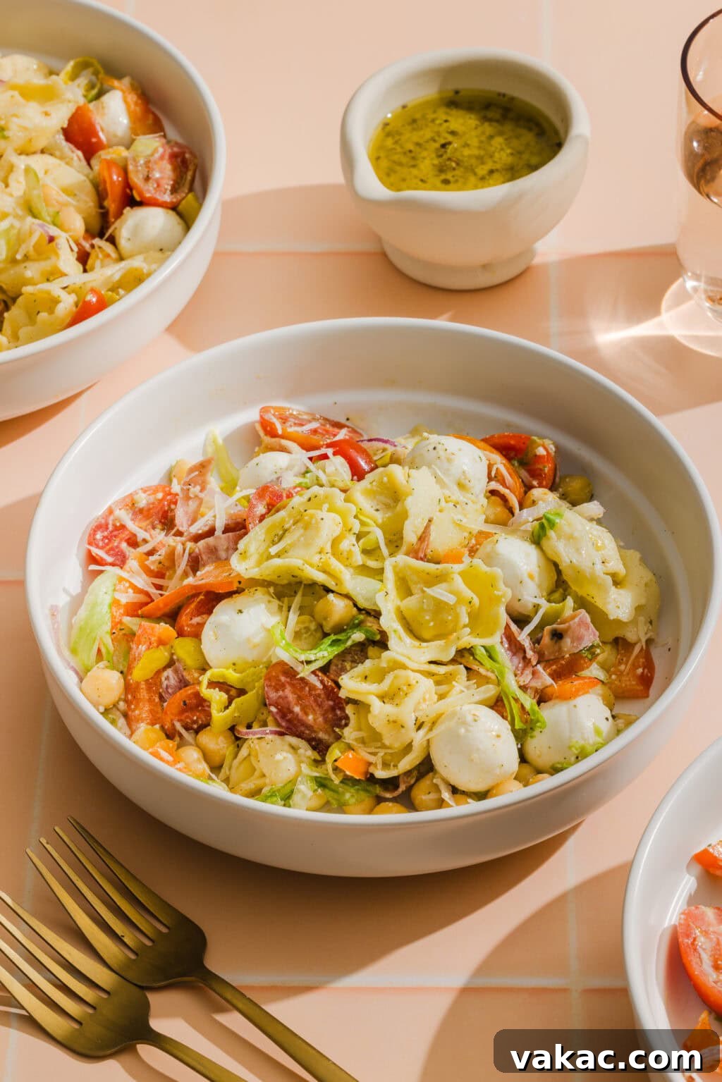 A bowl of tortellini pasta salad with mozzarella balls, cherry tomatoes, lettuce, and sliced salami, served with a side of green dressing. Gold forks and other salad bowls are visible on a peach-colored tiled surface.