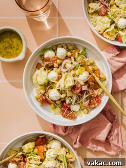 Three bowls of pasta salad with mozzarella balls, chickpeas, cherry tomatoes, lettuce, and shredded cheese on a peach tiled surface, next to a cup of water, a small dish of dressing, and a pink napkin.