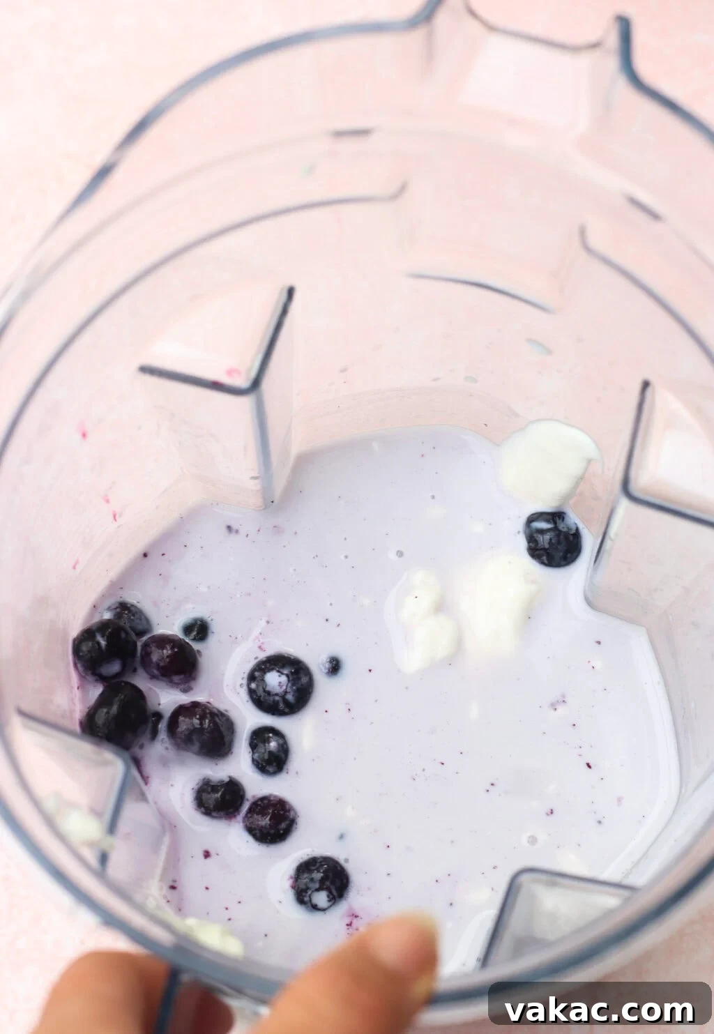 High-Protein Blueberry Chia Pudding (Blended for Creaminess) 4 A top-down view of a blender containing a small amount of milk or yogurt, several blueberries, and a few dollops of yogurt. A hand is holding the edge of the blender.