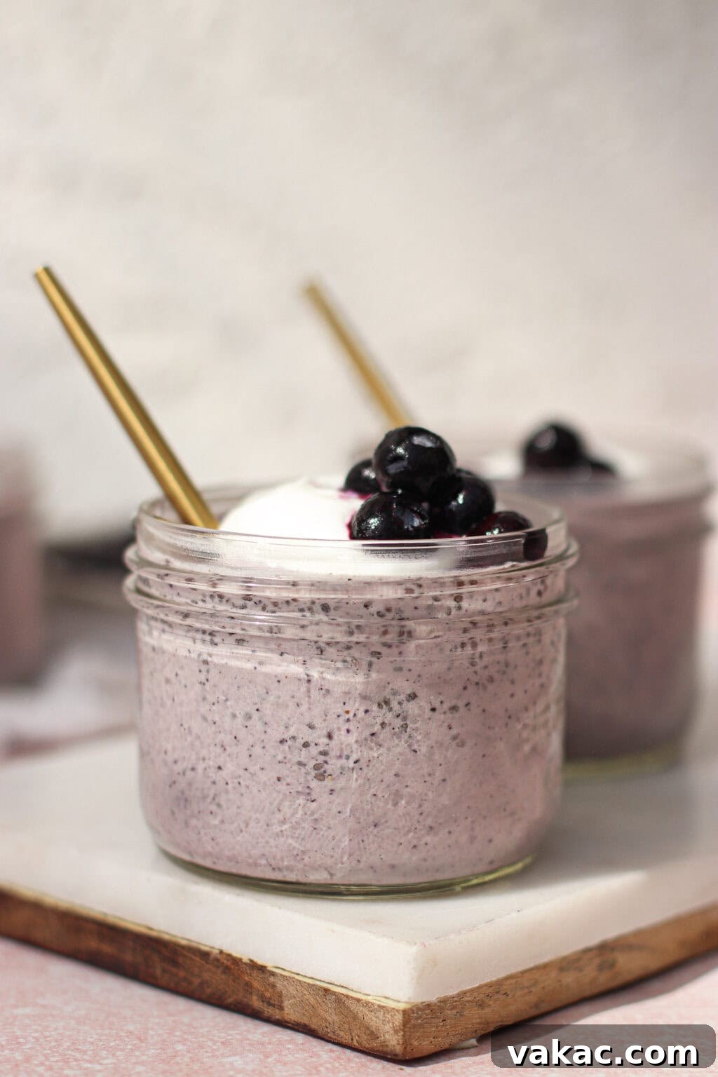 High-Protein Blueberry Chia Pudding (Blended for Creaminess) 7 Two glass jars filled with creamy purple smoothie, topped with whipped cream and blueberries, each with a gold straw, placed on a white marble slab.