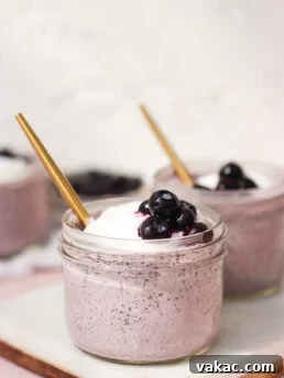 A glass jar filled with a creamy purple smoothie, topped with whipped cream and blueberries, with a gold straw. Another similar jar is in the background, both placed on a white surface.