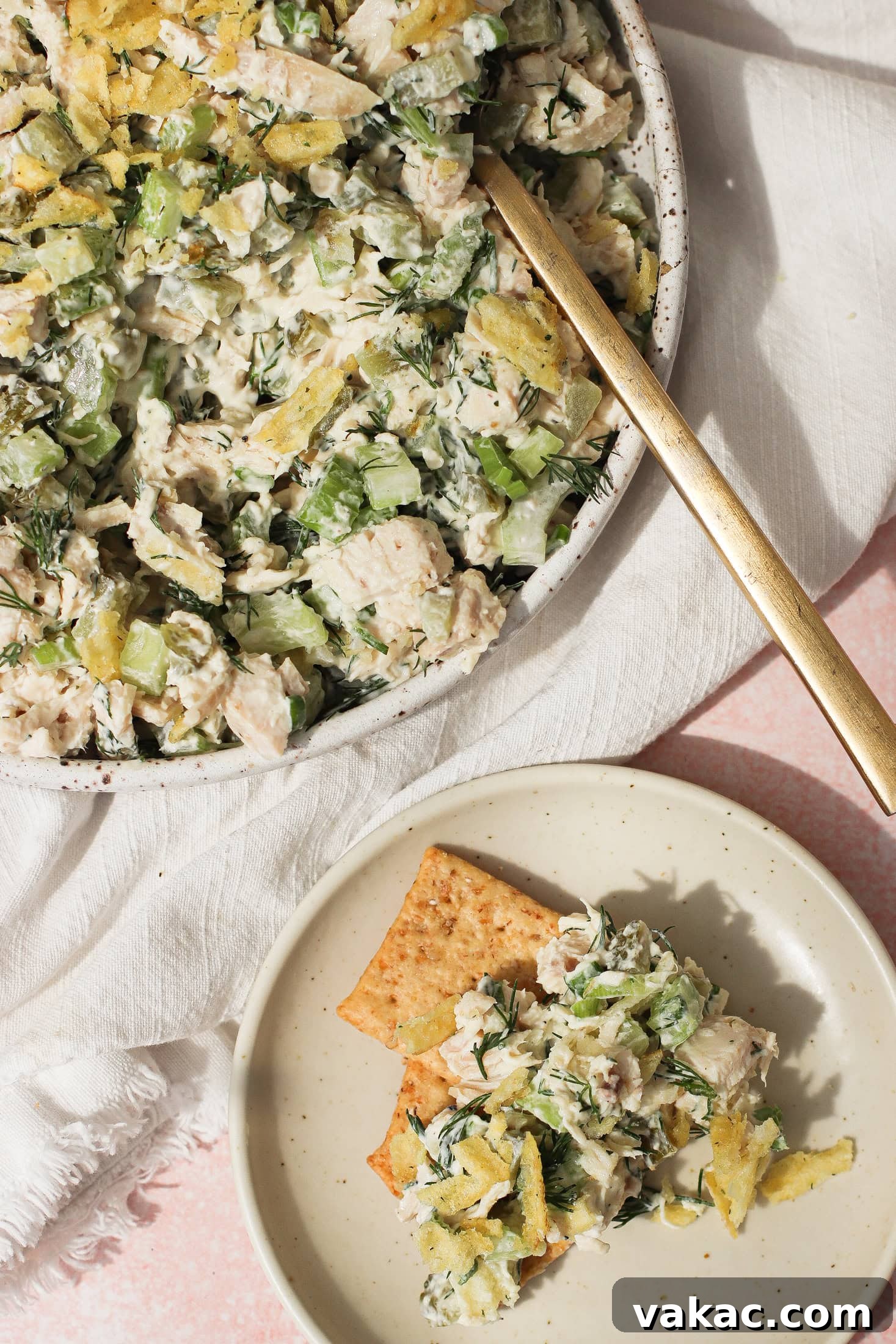 Chopped Dill Pickle Chicken Salad - High-Protein Recipe 2 A bowl of chicken salad with celery, herbs, and crushed potato chips, served with a gold fork. A small plate nearby holds crackers topped with the same chicken salad mixture.