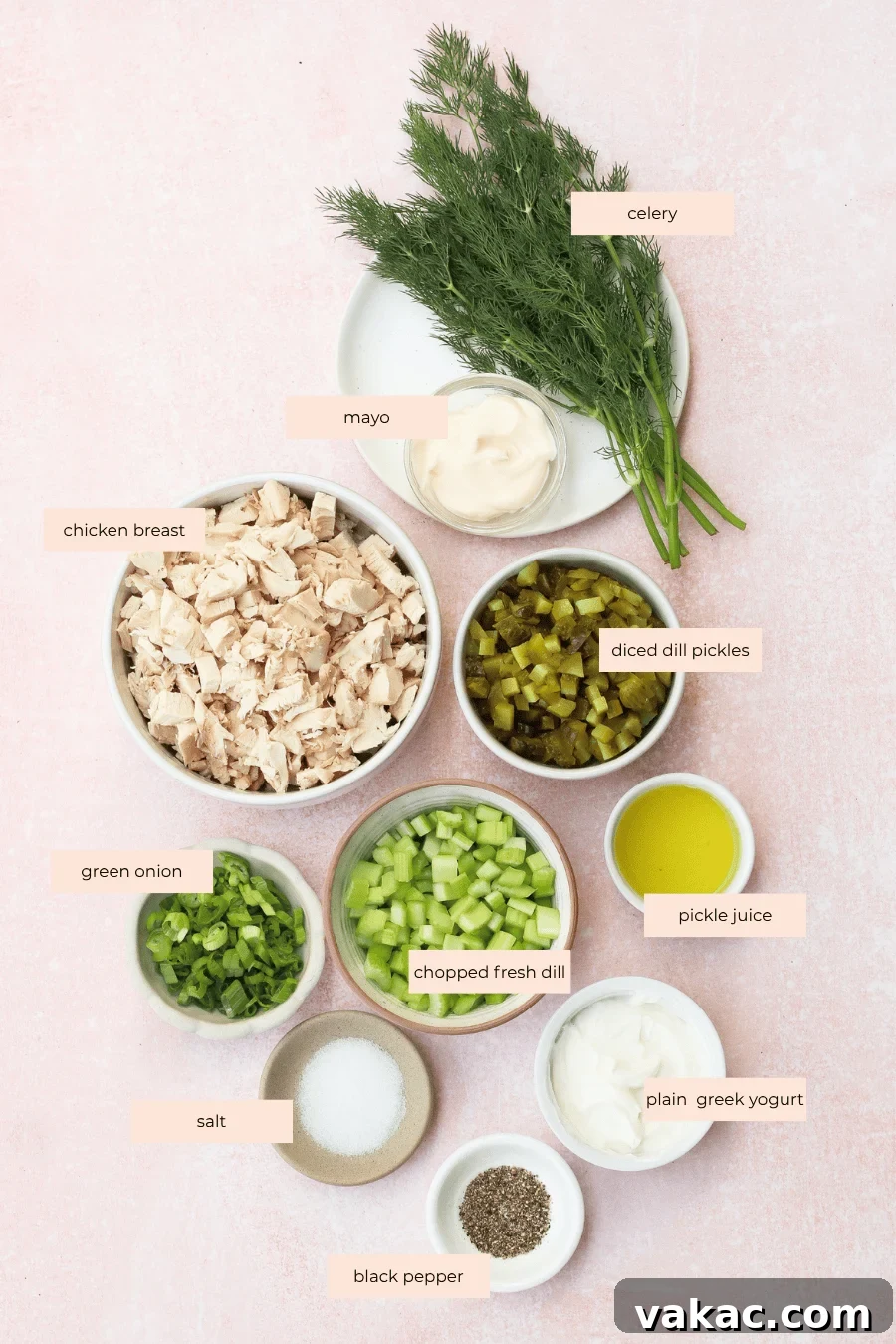 Chopped Dill Pickle Chicken Salad - High-Protein Recipe 3 Ingredients for a dill pickle chicken salad laid out on a light pink surface, including chopped chicken breast, green onion, celery, dill, diced pickles, mayo, salt, black pepper, yogurt, pickle juice, and herbs. Labels identify each ingredient.