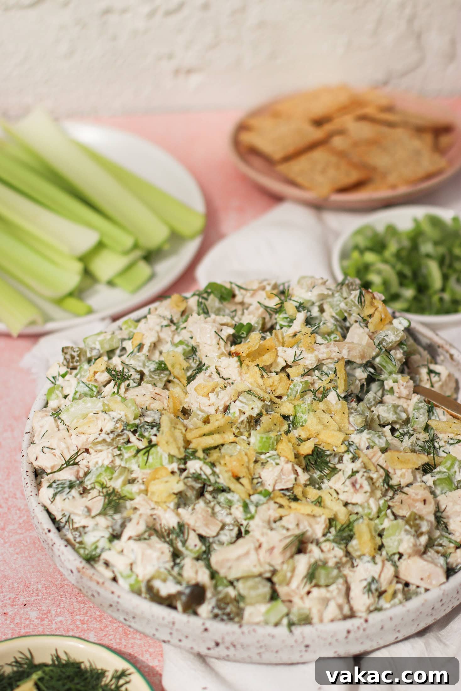Chopped Dill Pickle Chicken Salad - High-Protein Recipe 4 A bowl of creamy chicken salad garnished with crumbled potato chips sits on a table, surrounded by plates of celery sticks, crackers, and sliced green onions.