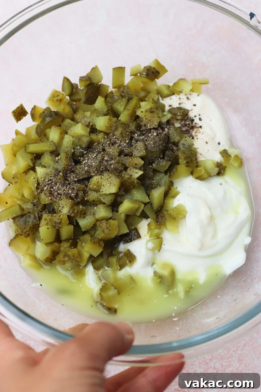Chopped Dill Pickle Chicken Salad - High-Protein Recipe 5 A clear glass bowl containing chopped pickles, yogurt or sour cream, pickle juice, and ground black pepper, held by a hand against a light pink background.