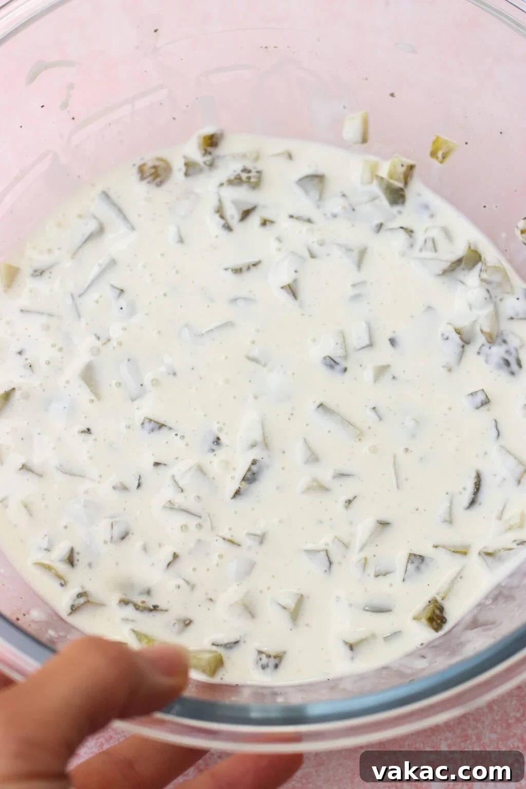 Chopped Dill Pickle Chicken Salad - High-Protein Recipe 6 A hand holds a glass bowl filled with a creamy white mixture containing chopped pickles and specks of black pepper.