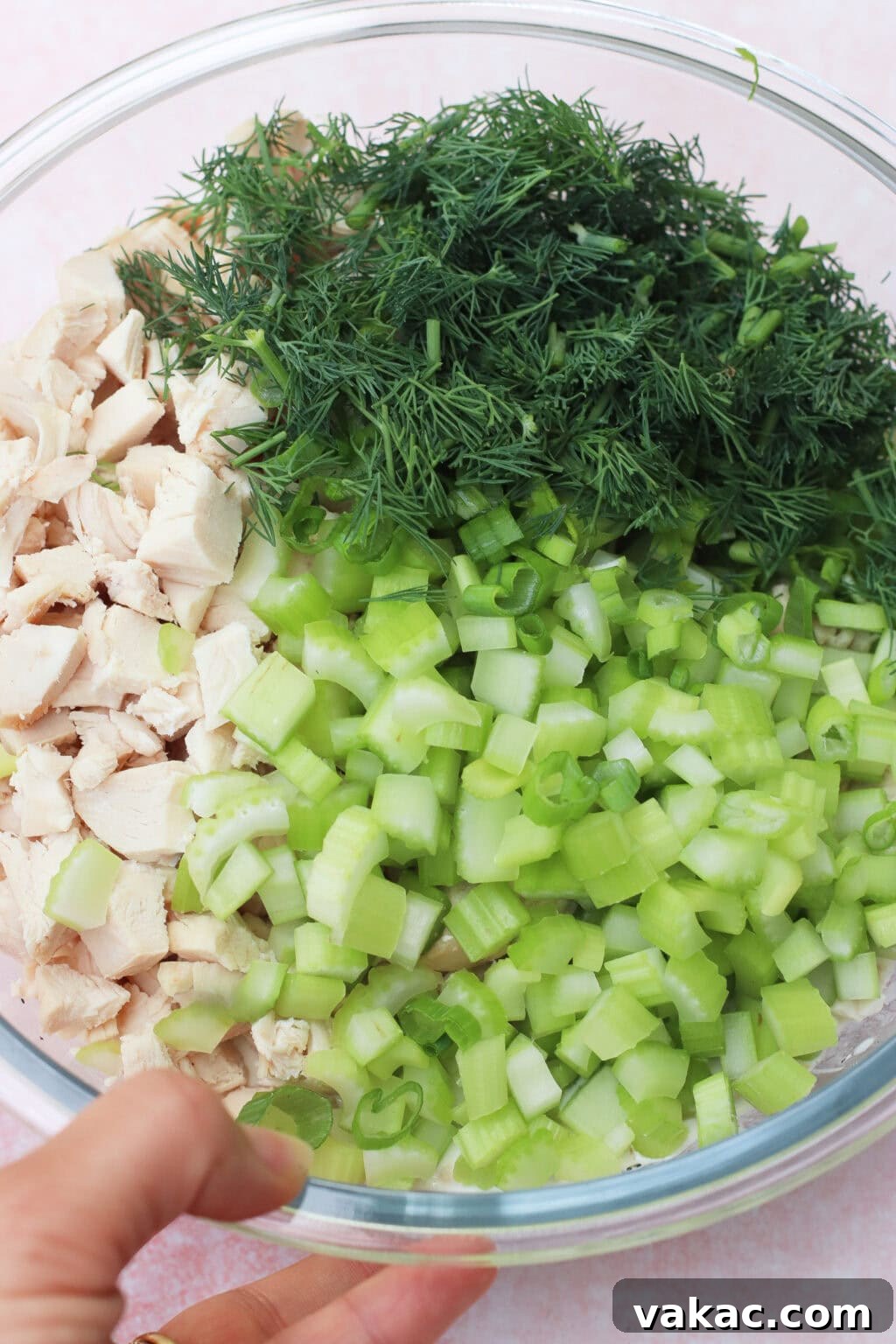 Chopped Dill Pickle Chicken Salad - High-Protein Recipe 7 A clear glass bowl containing chopped cooked chicken, fresh dill, chopped celery, and sliced green onions, with a hand holding the side of the bowl.