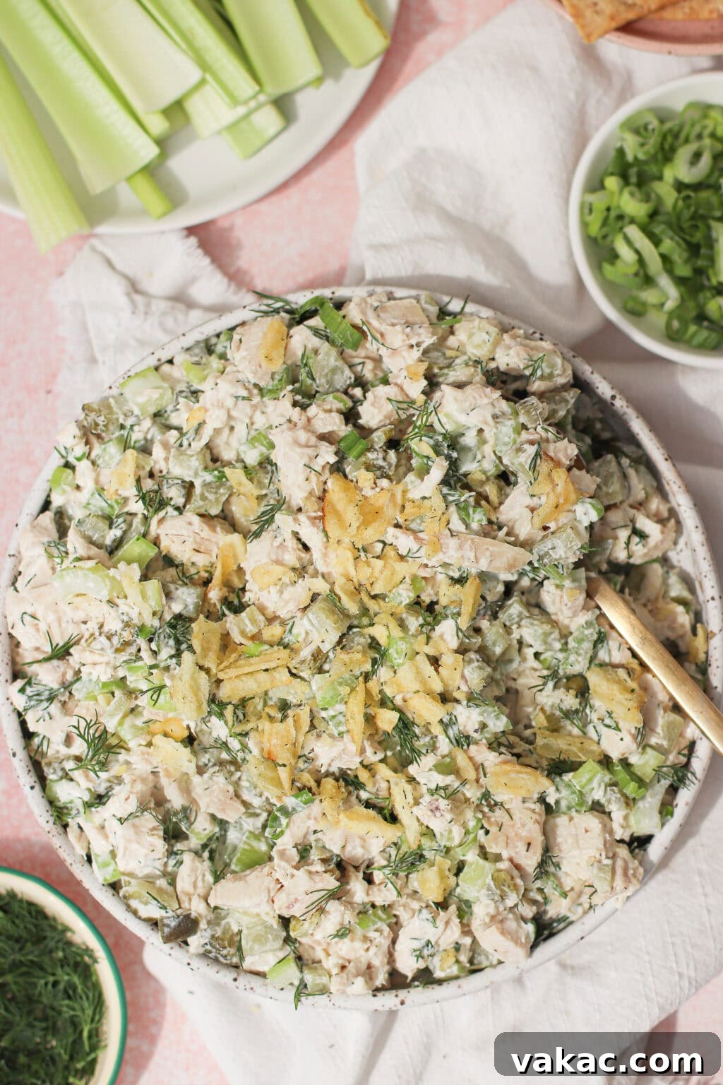 Chopped Dill Pickle Chicken Salad - High-Protein Recipe 8 A bowl of chicken salad mixed with herbs and topped with crushed potato chips, surrounded by celery sticks, sliced green onions, and dill on a light pink surface.