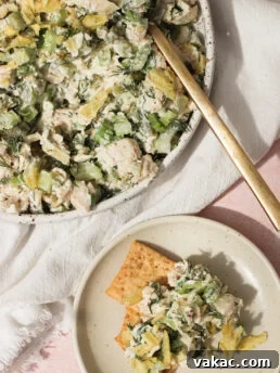 A bowl of chicken salad with celery, herbs, and crushed potato chips, served with a gold fork. A small plate nearby holds crackers topped with the same chicken salad mixture.