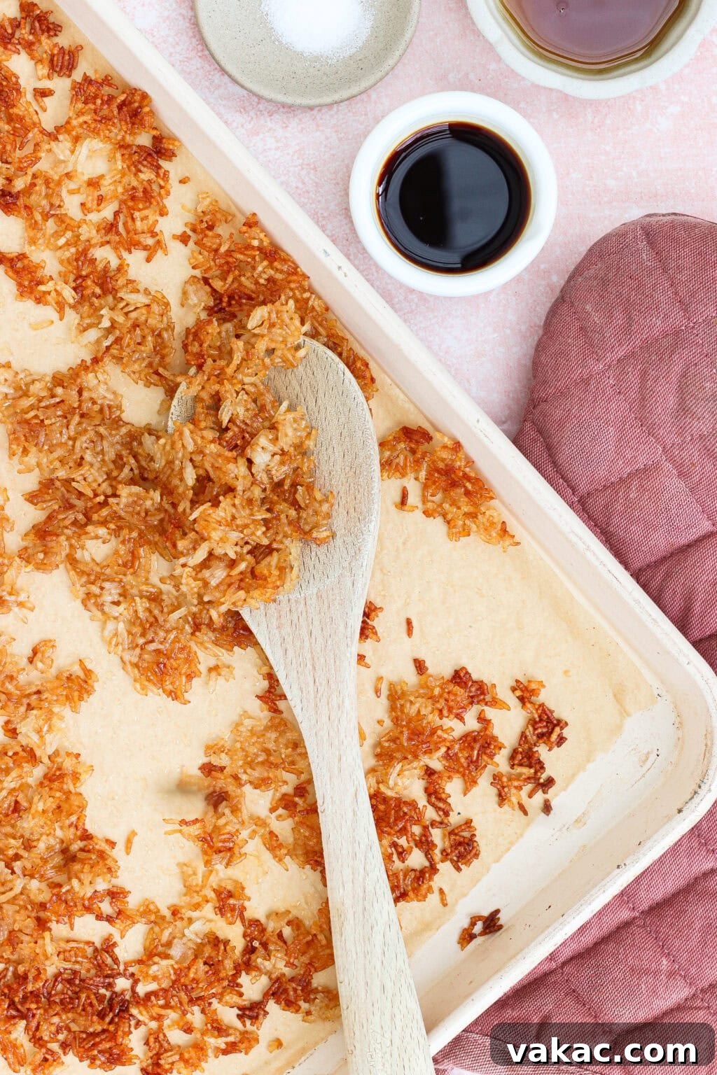 Crispy Sheet Pan Rice for Salads, Bowls and Toppings 2 A baking tray with toasted coconut flakes being stirred with a wooden spoon, surrounded by a pink oven mitt, a bowl of salt, and two small bowls with dark liquid.
