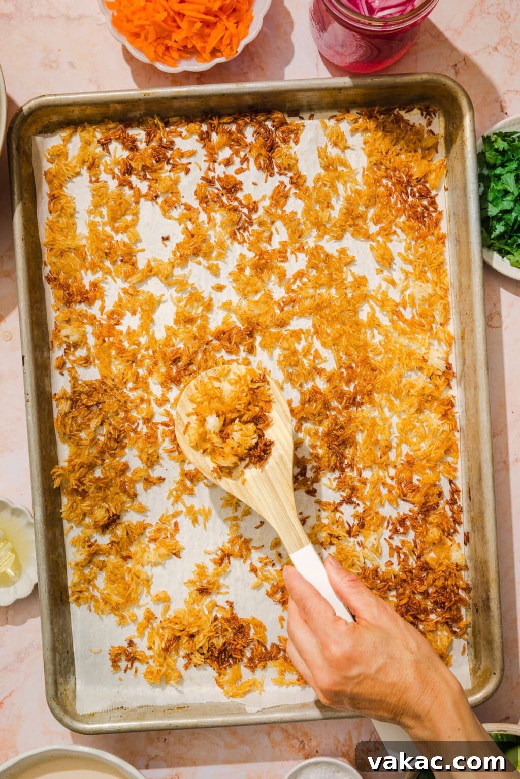 Crispy Sheet Pan Rice for Salads, Bowls and Toppings 7 A hand uses a wooden spoon to scoop crispy golden-brown fried shallots from a parchment-lined baking tray, surrounded by small bowls of colorful ingredients.
