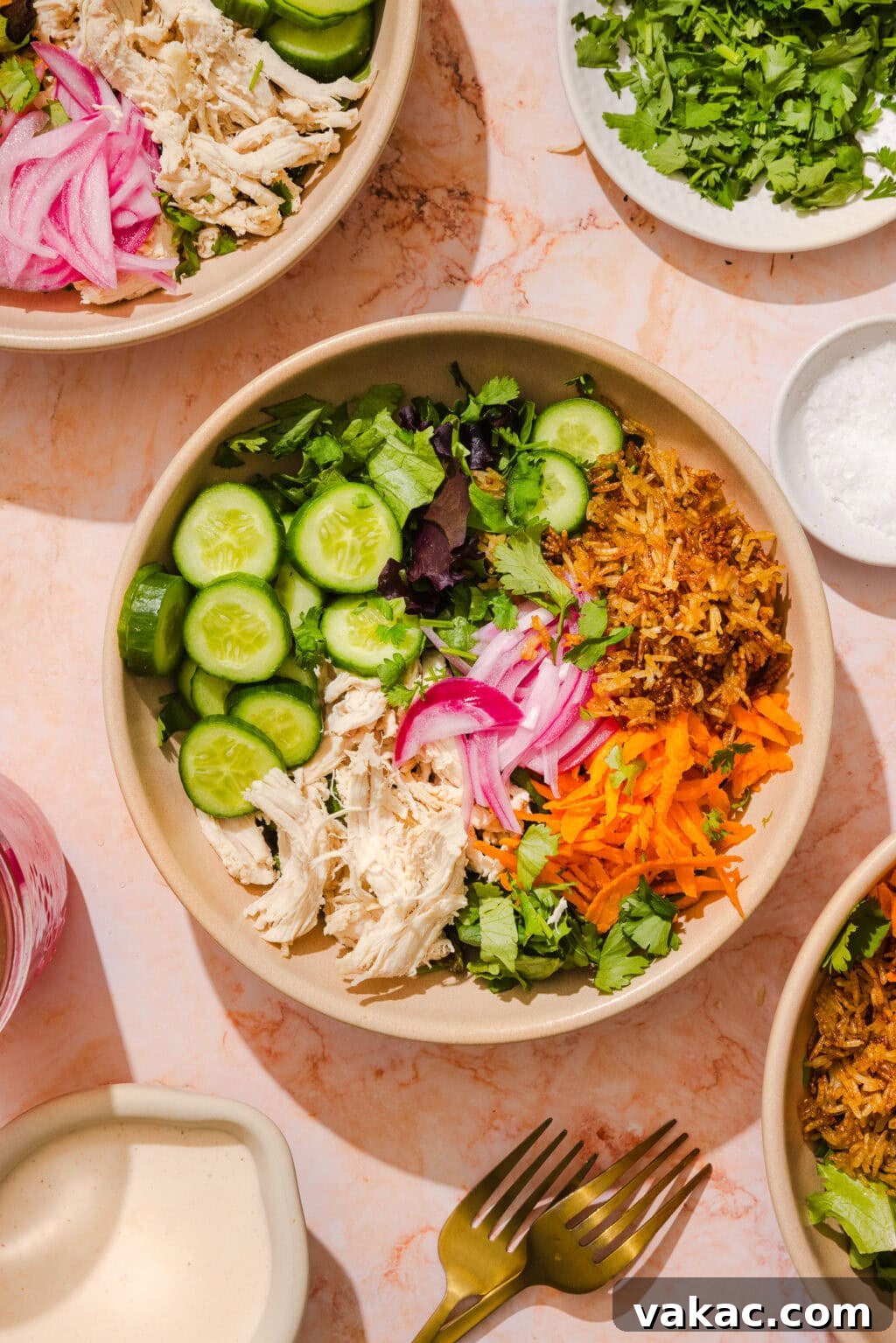 Crispy Sheet Pan Rice for Salads, Bowls and Toppings 8 A colorful salad bowl with shredded chicken, sliced cucumbers, pickled onions, shredded carrots, greens, cilantro, and crispy fried onions, surrounded by plates of herbs, salt, a sauce, and two forks on a marble surface.