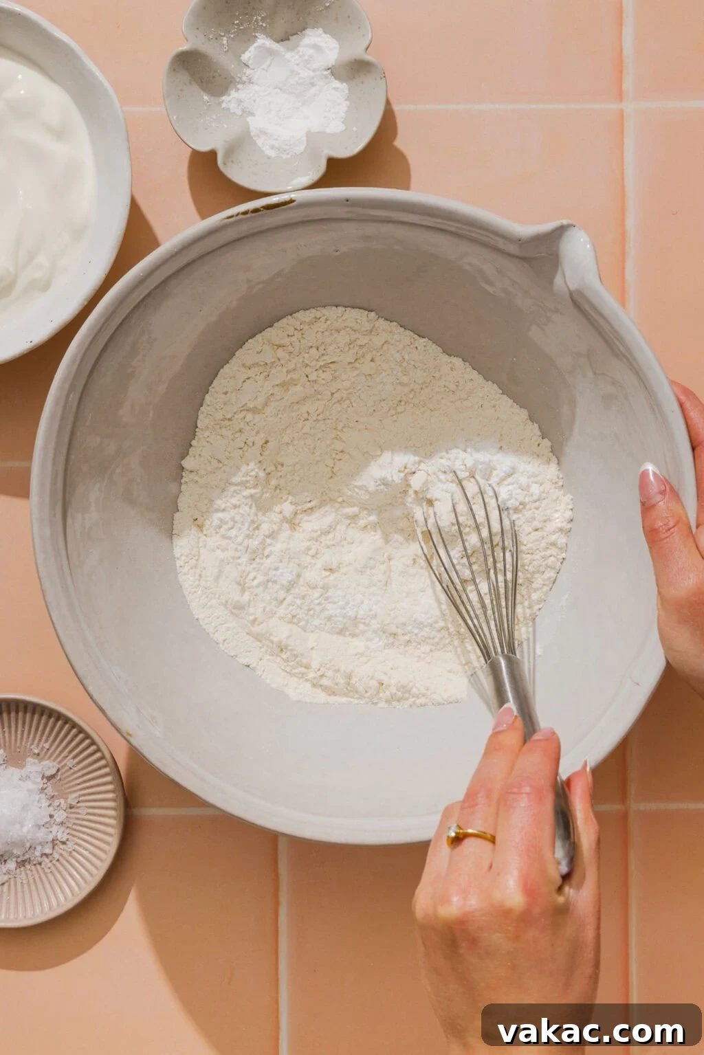 Protein-Packed Greek Yogurt Pizza Dough 6 A person whisks flour in a large bowl, surrounded by small dishes of baking powder, salt, and a bowl of yogurt on a peach-colored tiled surface.