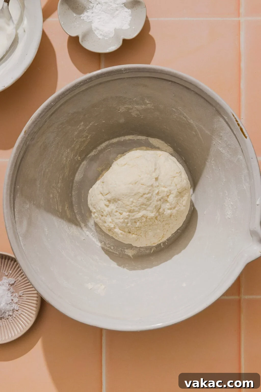 Protein-Packed Greek Yogurt Pizza Dough 7 A ball of dough sits in a large mixing bowl on a peach-colored tiled surface, surrounded by small dishes containing flour and other baking ingredients.