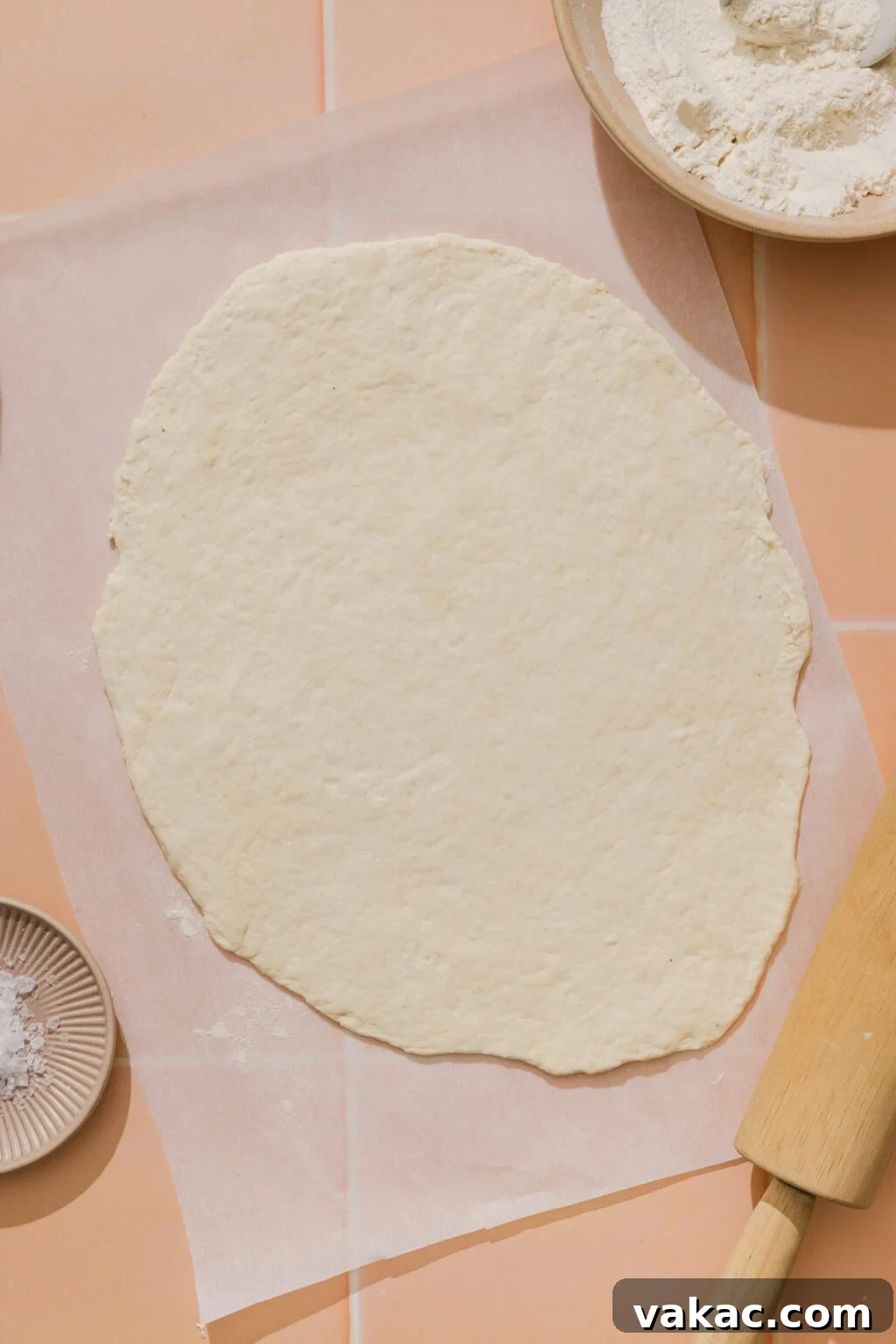 Protein-Packed Greek Yogurt Pizza Dough 8 Rolled out pizza dough on parchment paper, surrounded by a bowl of flour, a rolling pin, and a small dish of salt on a peach-colored tiled surface.