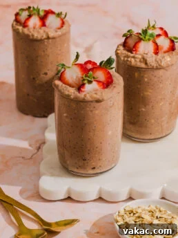 Three glasses of chocolate chia pudding topped with sliced strawberries are placed on a white tray, with two gold spoons and a small bowl of oats nearby on a pink marble surface.