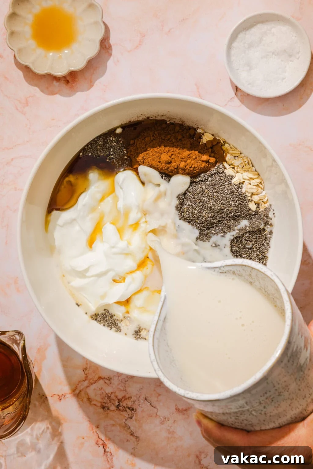 Protein-Packed Chocolate Overnight Oats with Greek Yogurt 5 A hand pours milk into a bowl containing yogurt, oats, chia seeds, cinnamon, and honey, with small dishes of honey and salt nearby on a marble surface.