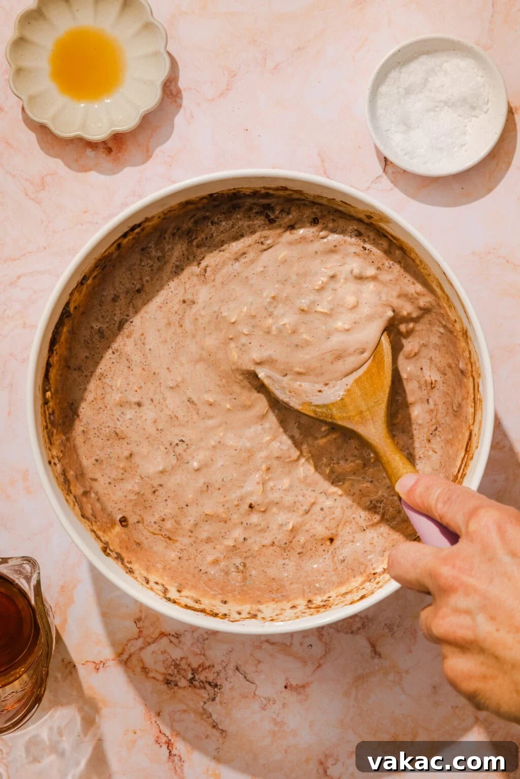Protein-Packed Chocolate Overnight Oats with Greek Yogurt 6 A hand stirs a bowl of chocolate batter with a wooden spoon on a marble surface; small dishes with salt and liquid ingredients are nearby.