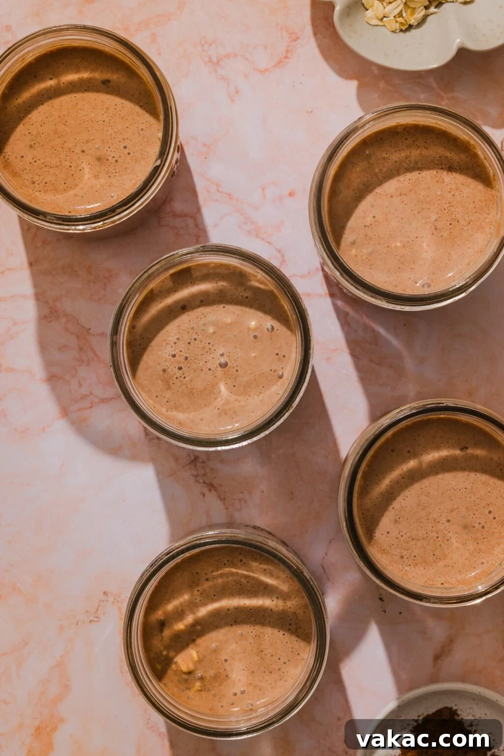 Protein-Packed Chocolate Overnight Oats with Greek Yogurt 7 Five glass jars filled with chocolate smoothie are arranged on a light marbled surface. The sunlight casts long shadows from the jars. Some oats and cocoa powder are visible in small bowls nearby.