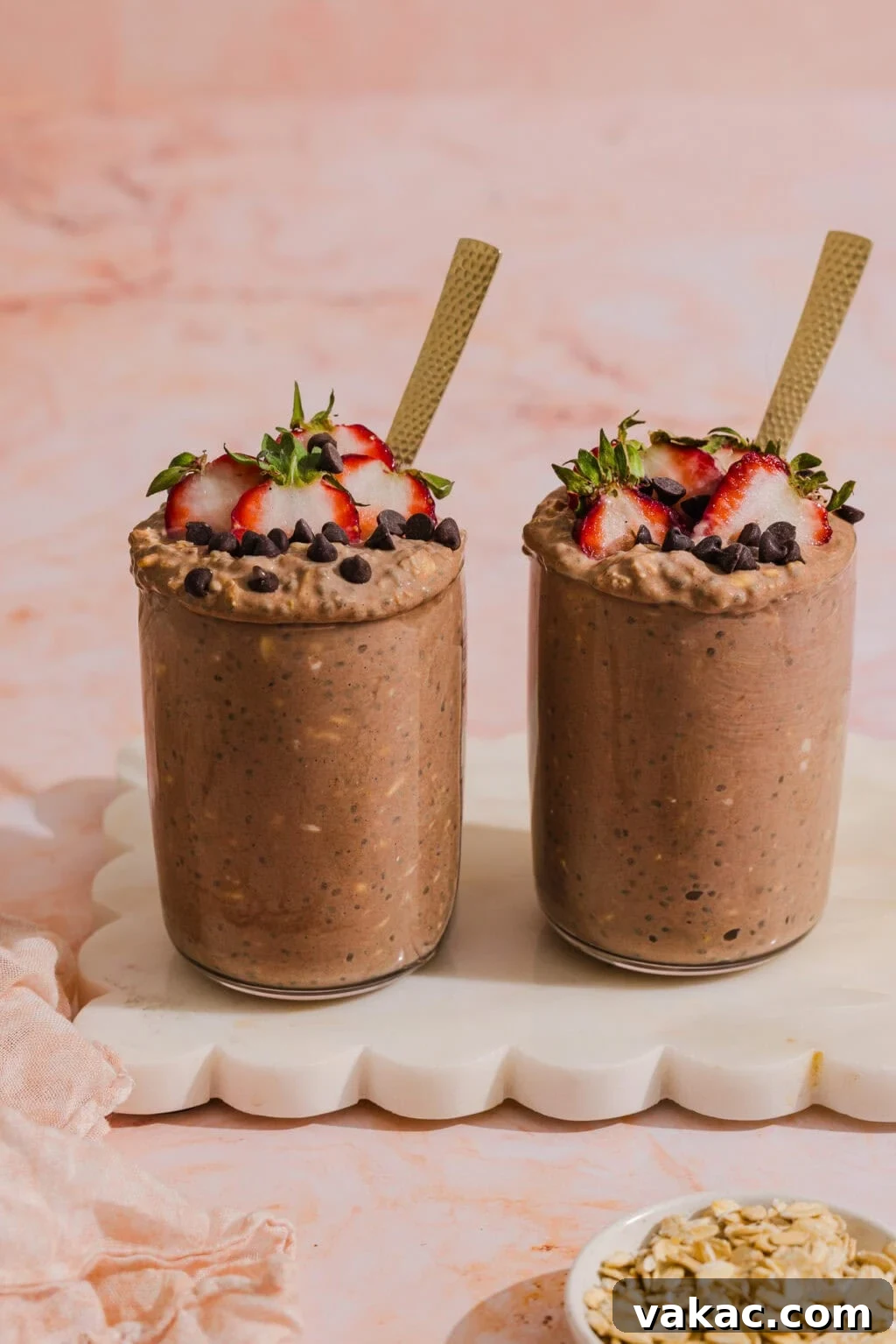 Protein-Packed Chocolate Overnight Oats with Greek Yogurt 9 Two glasses filled with chocolate overnight oats, topped with sliced strawberries and chocolate chips, each with a gold spoon, sit on a white marble tray. A small dish of oats is in the foreground on a pink surface.