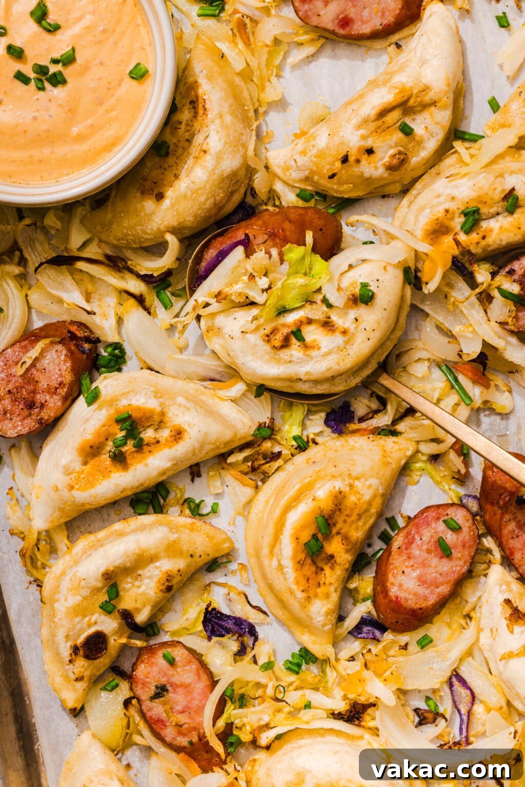 Sheet Pan Kielbasa and Pierogies from Frozen to Crispy 2 Pierogies with grilled sausage, sautéed onions, cabbage, and chives arranged on a tray, with a bowl of creamy dipping sauce in the corner.