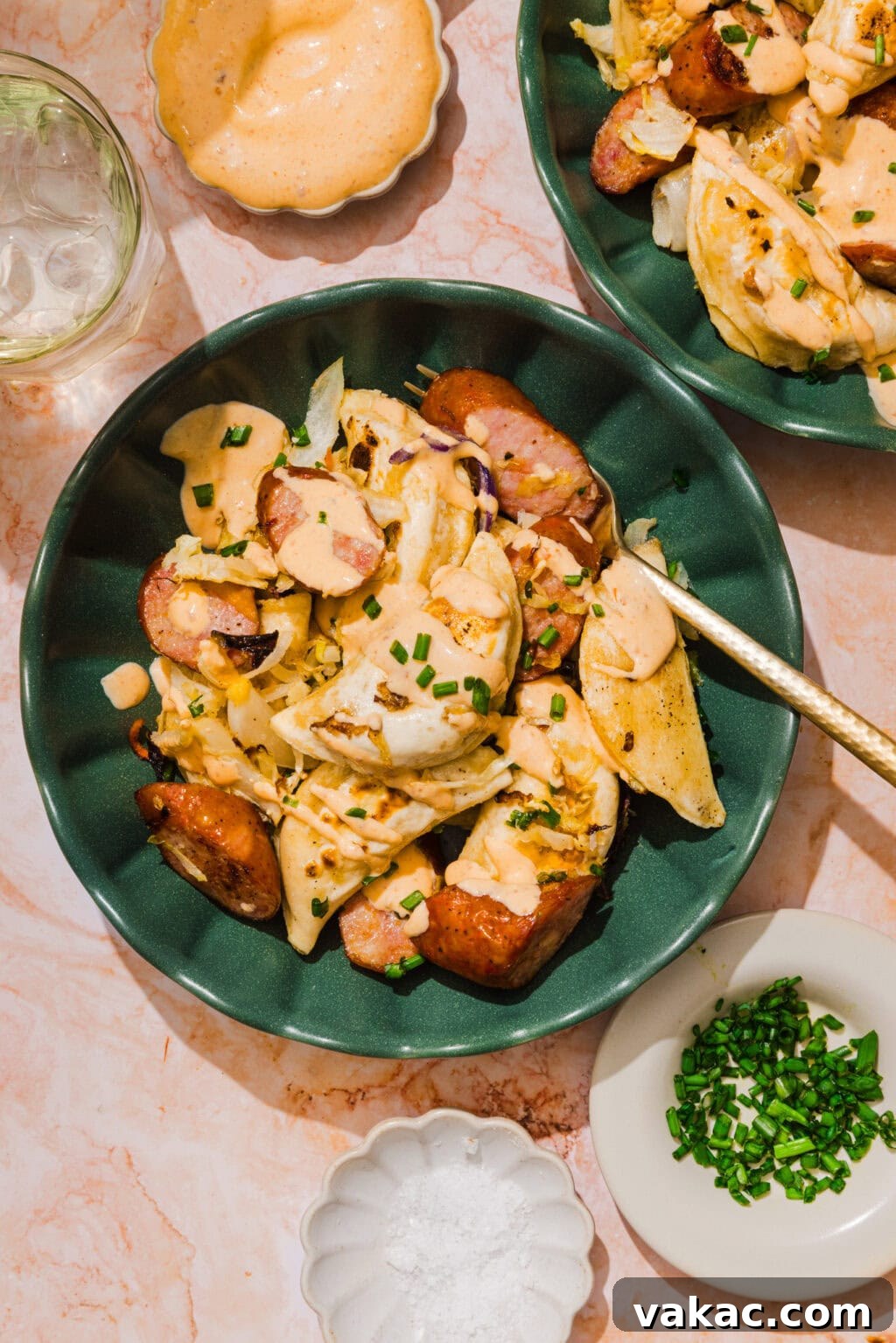 Sheet Pan Kielbasa and Pierogies from Frozen to Crispy 4 A green bowl with pierogi, sliced sausage, onions, creamy sauce, and chopped chives on top, with a fork. Surrounding dishes hold chives, salt, extra sauce, and a glass of water on a pink marble surface.