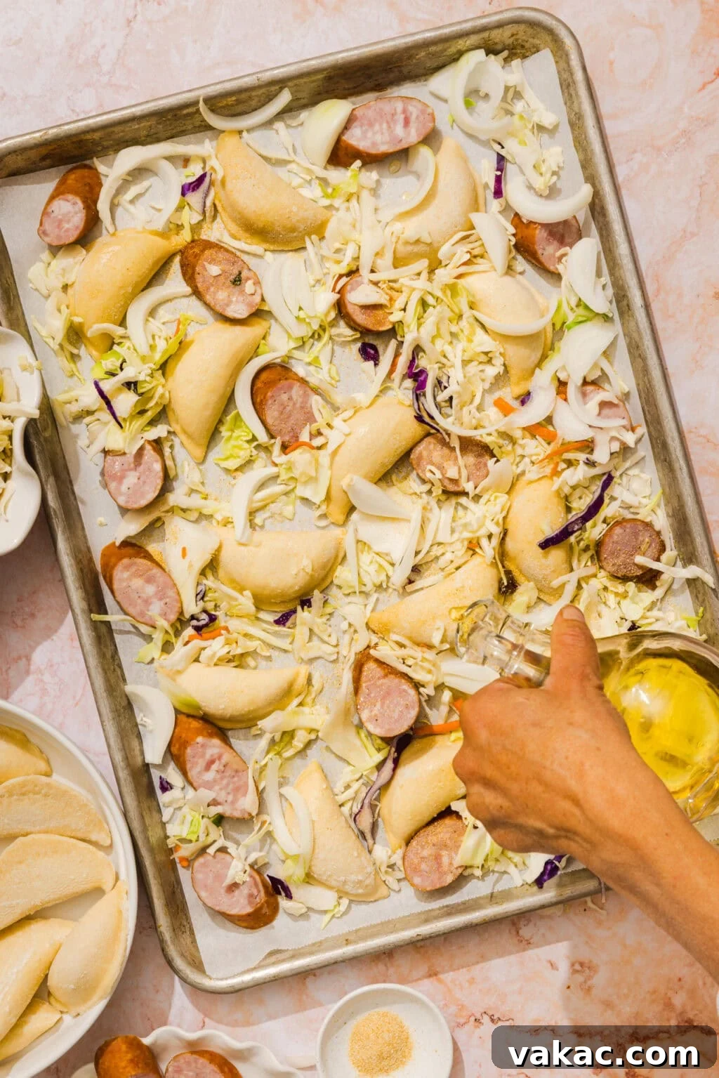 Sheet Pan Kielbasa and Pierogies from Frozen to Crispy 5 A hand pours oil over a baking sheet with pierogies, sliced sausage, cabbage, and onion, all spread out and ready for roasting. A bowl of seasonings and a plate are visible nearby.