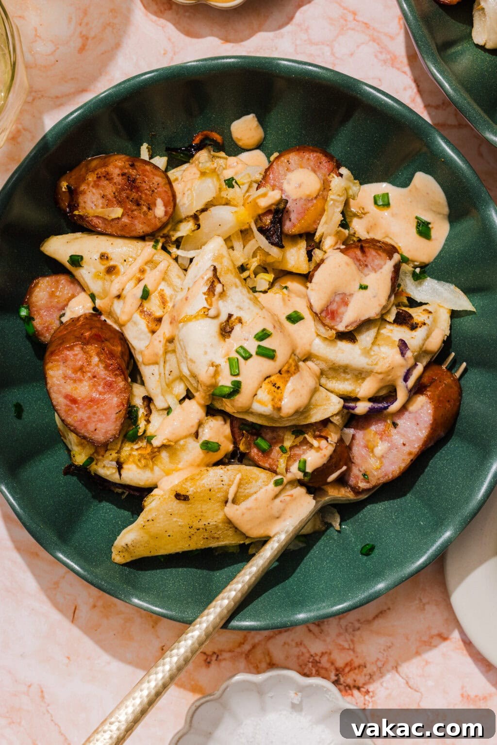 Sheet Pan Kielbasa and Pierogies from Frozen to Crispy 7 A green plate filled with pierogi, sliced sausage, sautéed onions, and creamy sauce, garnished with chopped chives. A fork rests on the plate.