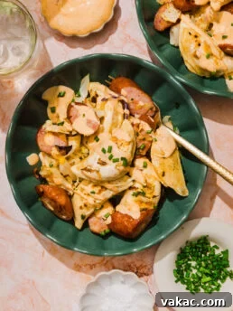 A green bowl with pierogi, sliced sausage, onions, creamy sauce, and chopped chives on top, with a fork. Surrounding dishes hold chives, salt, extra sauce, and a glass of water on a pink marble surface.